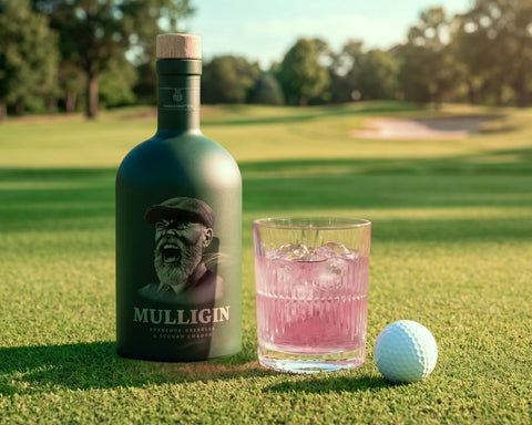 Bottle of Mulligin gin with a glass of pink cocktail and golf ball on a golf course.